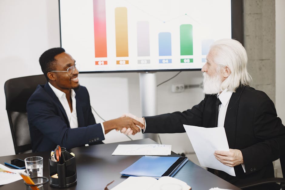 Two businessmen shaking hands in office setting with charts in background.